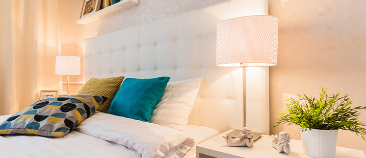 Romantic bedroom interior showing white faux leather headboard, white bedding with a aqua, yellow and mixed coloured dotted scatter cushions and matching white bedside lamps with glowing lights